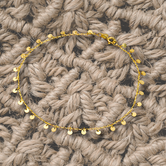 A gold-colored mini disc gypsy anklet laid out on a textured surface.