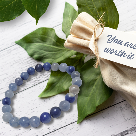 Blue and gray beaded bracelet on a white wooden surface with green leaves and a beige fabric pouch.Blue Aventurine Tumblestone Bracelet