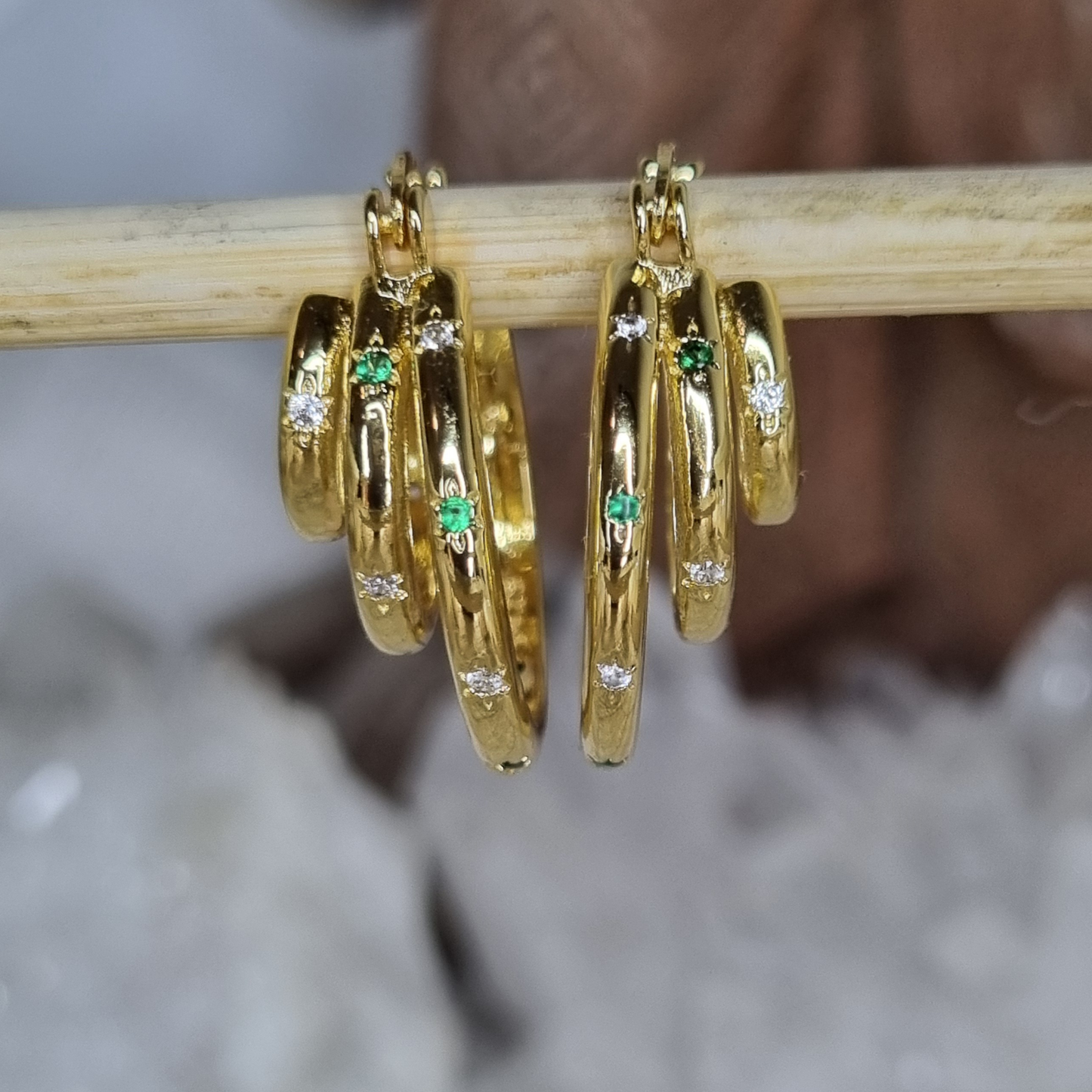 Gold hoop earrings with green gemstones and clear stones on a wooden stick.