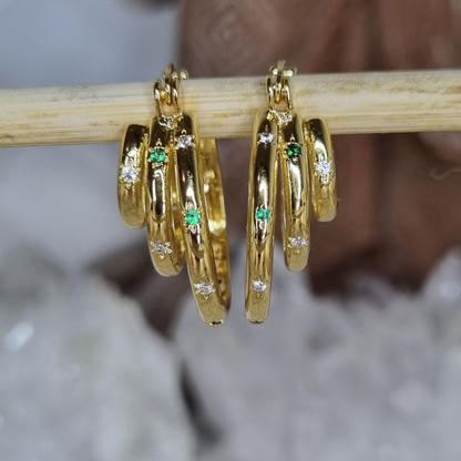 Gold hoop earrings with green gemstones and clear stones on a wooden stick.