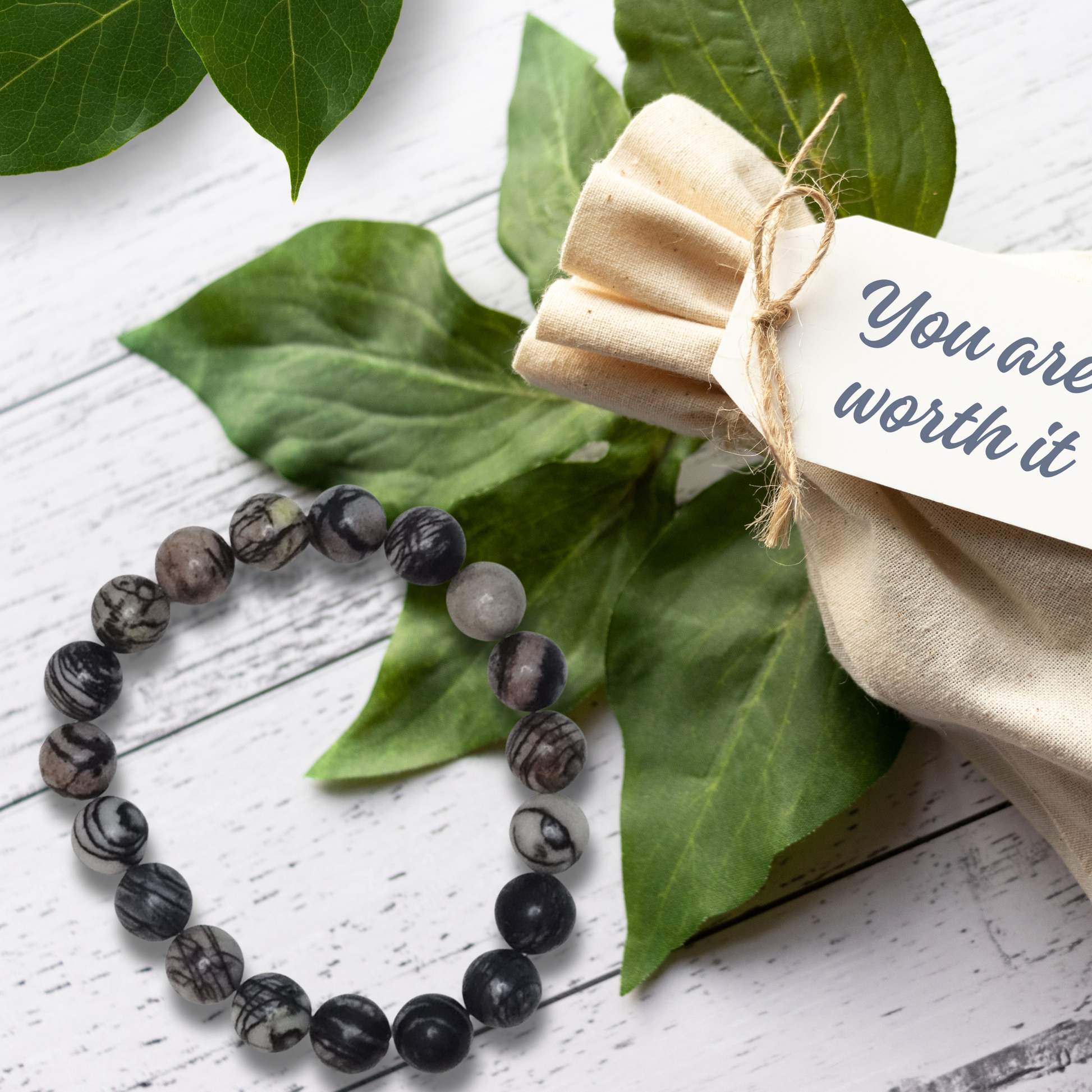 Black beaded bracelet on green leaves with a beige fabric pouch and tag.