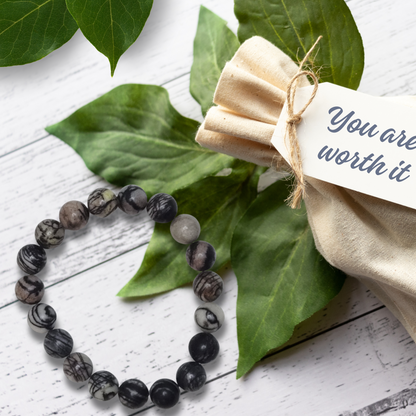 Black beaded bracelet on green leaves with a beige fabric pouch and tag.