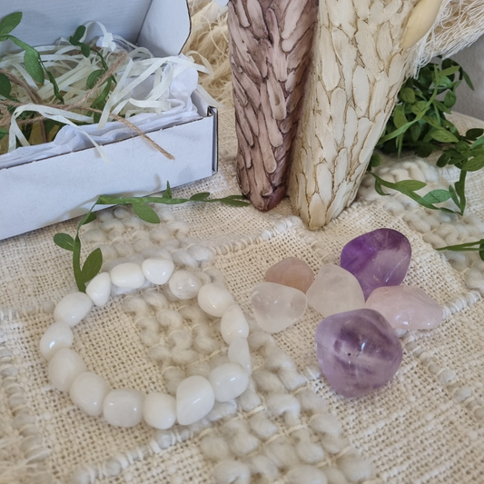 A white jade bracelet along with amethyst, rose quartz, and clear tumblestones displayed on a textured surface with a gift box and decorative elements in the background.