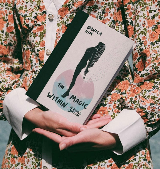 A person holding a hardcover guided journal titled 'The Magic Within' by Danica Gim, with illustrations and text on the cover.